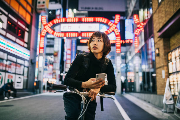 Woman Working as Bike Courier Young Asian Woman working for an food delivery app company. Using Smartphone to navigate in the big city, fluorescent light photos stock pictures, royalty-free photos & images