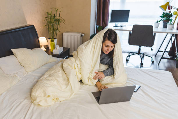 woman working in bed on laptop covered with blanket stock photo