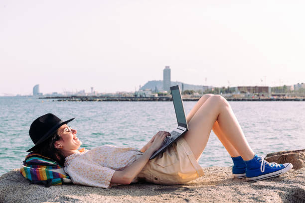 woman working lying down with computer by the sea young traveler woman working lying down with computer and backpack by the sea next to the city, concept of digital nomad and blogger lifestyle , copy space for text nomadic people stock pictures, royalty-free photos & images