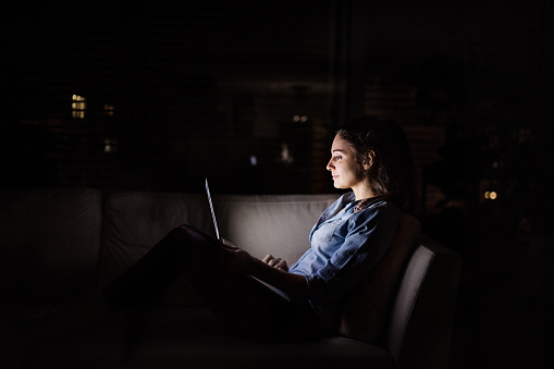 A woman sitting on a sofa at home at night, working on a laptop.