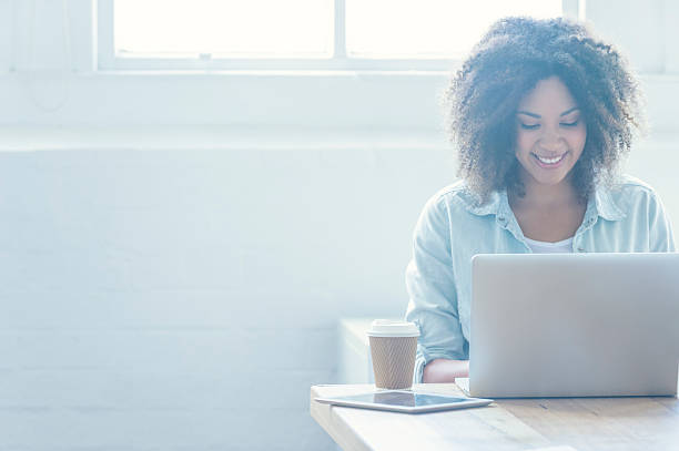 Woman working on a laptop. Woman working on a laptop. She is of African descent and has ah afro. There is also a digital tablet and coffee on the table. There is a large window behind her. Copy space job search photos stock pictures, royalty-free photos & images