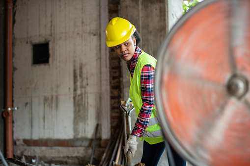 Female construction worker wearing reflective clothing and hardhat loading the concrete mixer using shovel at building site. Woman worker manually loading the cement mixer machine at construction site.