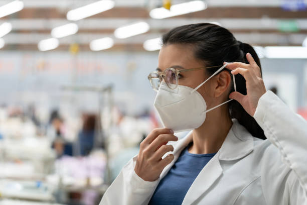 mujer trabajando en fábrica textil durante la pandemia covid-19 y arreglando su mascarilla - n95 mask fotografías e imágenes de stock