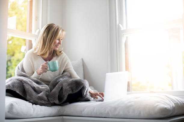 Woman wrapped in blanket using laptop at home Mature woman holding coffee cup while working on laptop. Smiling female is using technology on window seat. She is wearing casuals at home. alcove window seat stock pictures, royalty-free photos & images