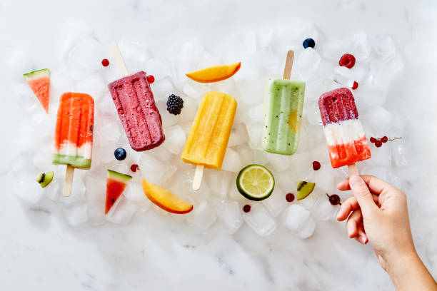 A woman's hand holds an ice cream popsicle against a background of ice cubes with ice cream and a piece of fruit. Flat lay In the girl's hand, a healthy berry ice cream lolly . On the ice cubes, a different ice cream with pieces of fruit and fresh berries. Cold dessert. Flat lay flavored ice stock pictures, royalty-free photos & images