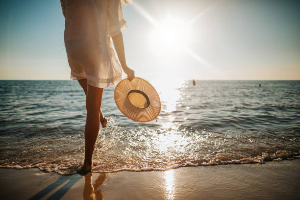 Woman's legs splashing water on the beach Close-up of young woman in white sun dress and with hat in hand walking alone on sandy beach at summer sunset, splashing water in sea shallow walking on beach stock pictures, royalty-free photos & images