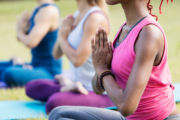 Women outdoors doing yoga, lotus pose with prayer hands Cropped view of a multi-ethnic group of three women taking a yoga class. They are outdoors, on exercise mats on the grass, in the lotus position with prayer hands. The focus is on the African-American woman in the foreground. asian yoga pants stock pictures, royalty-free photos & images
