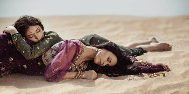 Women thirsty laying in a desert. Lost in desert durind sandshtorm Desert women lying on sand outdoors. Dehydration, overheating, thirst and heat stroke concept image with two sisters outdoors in the nature. Two arabian girls lost in desert during journey. Windy weather hot arabic girl stock pictures, royalty-free photos & images