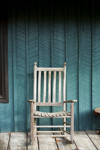 A single wooden rocking chair on a porch. The brown leaves on the floor indicate that it is autumn.