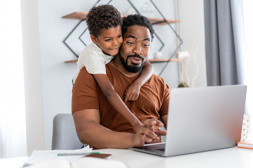 African American father using laptop and working at home while being distracted by his son.