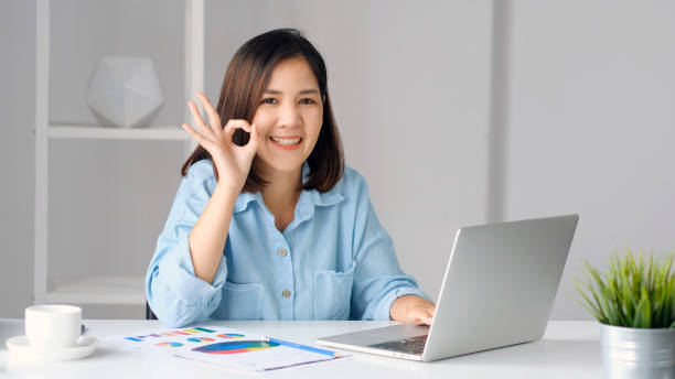 Work from home, Happy asian women working with laptop computer at home office and showing ok hand sign, Asia female with okay hand sign and smiling while woking at home office stock photo
