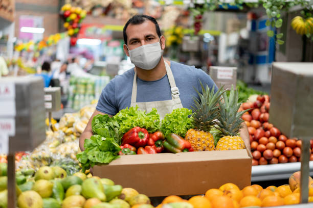 Worker at a food market restocking the shelves and wearing a facemask Latin American worker at a food market restocking the shelves and wearing a facemask while holding a box with fruits and vegetables - pandemic lifestyle concepts farmer's market stock pictures, royalty-free photos & images