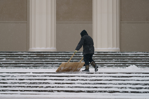 Moscow, Russia - January 26, 2022: Municipal city service cleans staircase from snow after blizzard in Moscow public park. Worker shoveling snow on sidewalk after snowfall. Working man. Janitor digging snow with shovel. Occupation