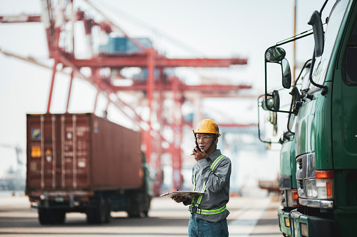 Dock worker standing against cargo containers having conversation with loading control room for shipping plans