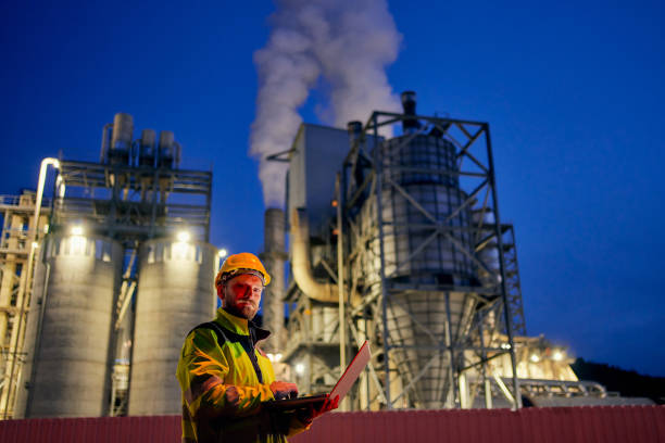 Worker with safety helmet and laptop working in factory during night shift Young worker working with a laptop in front of oil factory in the evening oil refinery factory stock pictures, royalty-free photos & images