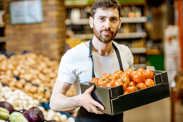 Worker with vegetables in the supermarket Portrait of a handsome shop worker or farmer holding box with fresh tomatoes in the vegetable department in the supermarket homegrown produce stock pictures, royalty-free photos & images