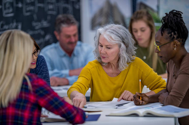 Working Together On A Project A group of university students are indoors in a study hall. They are having a group discussion. A Caucasian senior woman is reading out loud from a textbook. adult education stock pictures, royalty-free photos & images