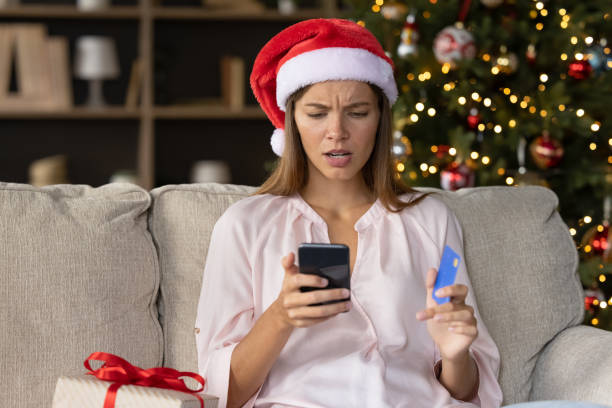 Worried concerned girl in Christmas Santa hat having problems Worried concerned girl in Christmas Santa hat having problems with payment by credit card online for New Year purchases, looking at smartphone screen with puzzled face. Scam, fraud concept scammer stock pictures, royalty-free photos & images