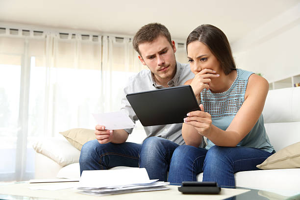 Worried couple checking bank account online Worried couple checking bank account trouble online in a tablet sitting on a couch in the living room at home common customer problems stock pictures, royalty-free photos & images