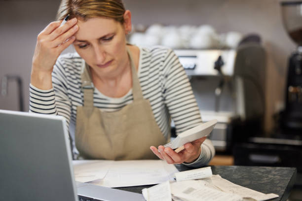 Worried Female Owner Of Coffee Shop In Financial Difficulty Looking Through Bills Using Laptop And Calculator stock photo