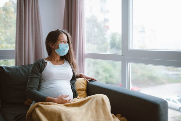 Worried pregnant woman with protective face mask at home. worried pregnant woman sitting at home with protective face mask and looking through the window. pregnant stock pictures, royalty-free photos & images
