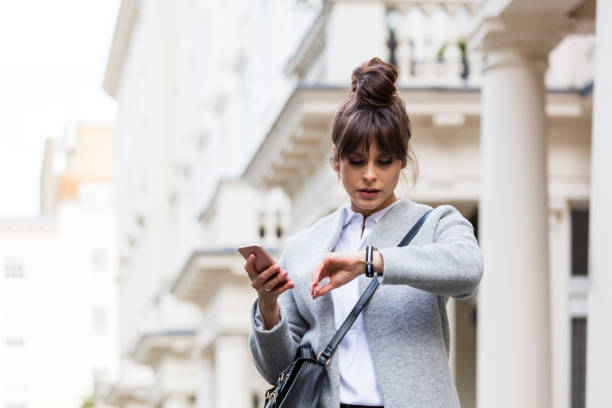 Worried woman standing with smart phone in front of city house Elegant worried woman standing in front of city house in London, holding smart phone in hands and checking time. Autumn season. beat the clock stock pictures, royalty-free photos & images