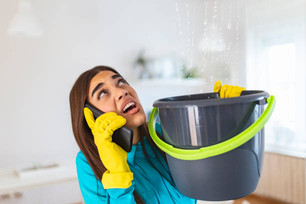 Worried Young woman Calling Plumber While Leakage Water Falling Into Bucket At Home stock photo