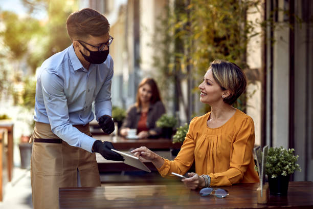 I would like to order this! Happy woman talking to a waiter who is wearing protective face mask while choosing something from a menu on touchpad in a cafe. restaurant stock pictures, royalty-free photos & images