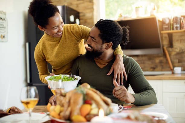 Would you like some salad, dear? Happy African American woman embracing her husband wile serving him salad during Thanksgiving lunch at dining table. mid adult couple stock pictures, royalty-free photos & images