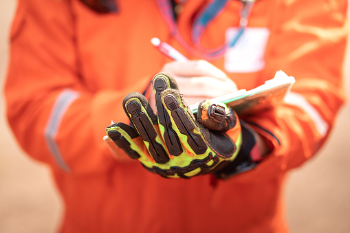 Close-up action of safety supervisor's hand which is weared an impact glove is holding a clipboard, and use the another hand writing something on paper during safety audit. Selective focus at glove.