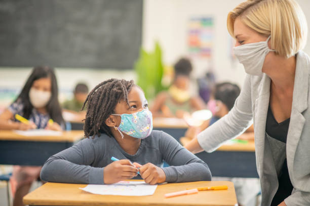 6 year old, African American student wearing a protective face mask in class Female student wearing a reusable face mask while sitting at her desk in class. lecture hall stock pictures, royalty-free photos & images