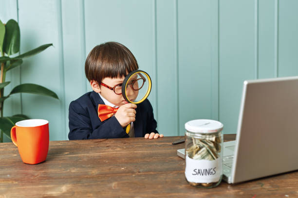 2-3 years old child wearing a suit like a businessman and he work in his office table. He looking to money with magnifying glass stock photo