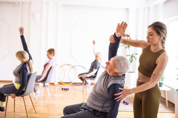 Yoga instructor assisting senior man during yoga class on chairs stock photo