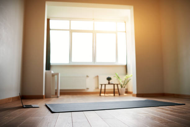 sala de yoga con gran ventana luminosa en piso moderno. Colchoneta de yoga y computadora en el piso, no hay sala de yoga para personas con una gran ventana luminosa en un piso moderno. Colchoneta de yoga y ordenador en el suelo, sin gente. tapete de casa fotos e imágenes de stock