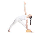 Sporty beautiful young beginning yoga female student standing in Utthita Trikonasana, Extended Triangle Pose with wooden block, studio full length profile view on white background, isolated