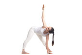 Sporty beautiful young woman in white sportswear standing in variation of Triangle yoga pose, studio full length three-quarters view on white background, isolated