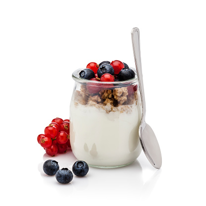 Front view of an open glass container filled with fresh yogurt with berries and granola isolated on white background. Some berries are out the container placed directly on the background. A spoon is beside the container. DSRL studio photo taken with Canon EOS 5D Mk II and Canon EF 100mm f/2.8L Macro IS USM
