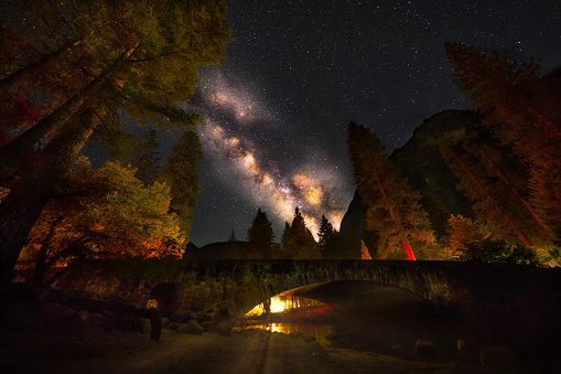 This image features the a magical view of Yosemite, over a tree lined bridge with a night sky illuminated with the milky way and shooting stars.
