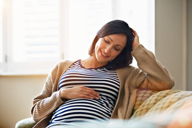 You'll be here any day now! Shot of a smiling pregnant woman sitting on her sofa holding her stomach pregnant stock pictures, royalty-free photos & images