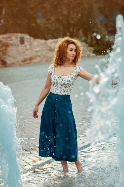 youn moroccan woman, with curly brown hair, wearing a jean skirt, playing with water at a fountain stock photo