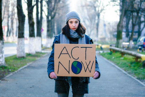Young activist holding sign protesting against climate change Portrait of a young teenage girl activist holding a sign protesting against climate change and global warming. She is standing outdoors on a city street and is holding signs with various messages, such as 'go green' and 'save our planet'. She is looking directly at the camera with a serious expression. Focus on the girl and her sign, with city street defocused beyond. Room for copy space. climate action stock pictures, royalty-free photos & images