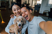 若い大人のアジアのカップルは、背景に家のインテリアと携帯電話から自分撮りを取って子犬を保持しています。家族の写真撮影犬のペットを持つ30年代の成熟した男女。- ハッピーグループ