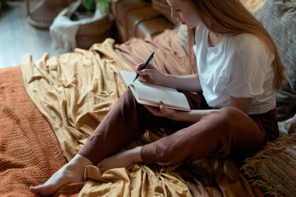 Young adult girl resting in home at cozy bedroom High angle view of thoughtful young adult girl writing notes in diary, sitting on bed, resting in home at cozy bedroom in boho style diary stock pictures, royalty-free photos & images