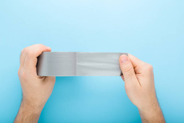 Young adult man hands stretching gray adhesive tape on light blue table background. Pastel color. Closeup. Point of view shot. Top down view. stock photo