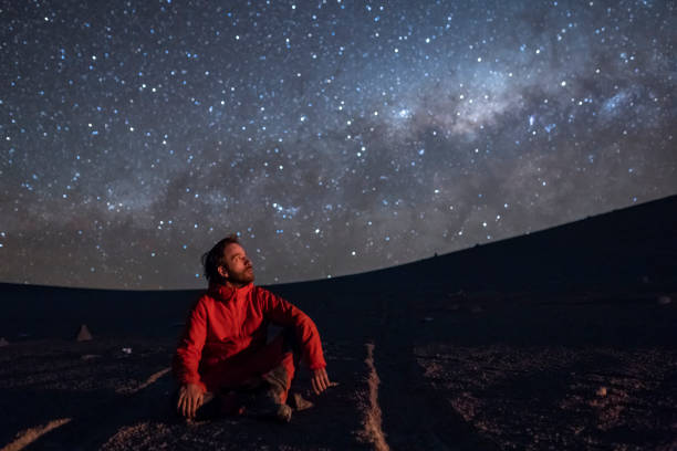 A young adult man seating and looking at the view of our Milky Way galactic core located in the constellation of Sagittarius, an amazing view at Atacama Desert. Chile Atacama Desert an amazing place in the southern hemisphere of the Earth, maybe the driest desert in the world is a combination of salt falts, salt lakes, amazing turquoise waters beaches, volcanoes and awesome landscape with the best night sky and an amazing place for looking for meteorites astronomy stock pictures, royalty-free photos & images