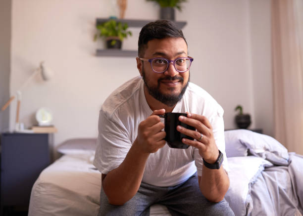 A young adult man smiles sitting on his bed drinking coffee in the morning stock photo