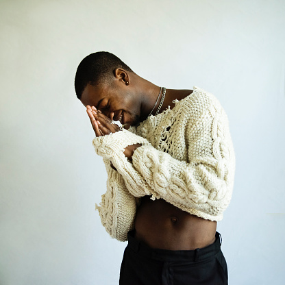 Young adult man with dark skin studio portrait. He is standing, with black pants and a custom made wool sweater, silver rings and necklace. Square indoors waist up studio shot in natural light with copy space.