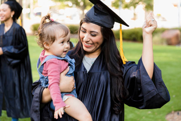 Young adult mom in cap and gown smiles at daughter The young adult mother, in her cap and gown, smiles as she teaches her baby daughter how to cheer. public universities in usa stock pictures, royalty-free photos & images