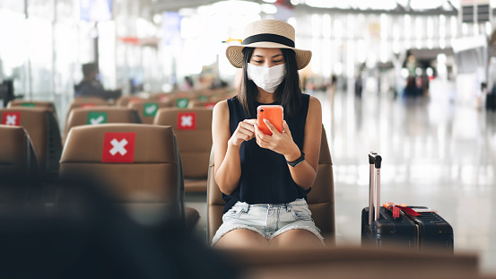 ヤングアダルト観光客の女性は社会的距離の椅子を備えた空港ターミナルでウイルス発生のためのマスクを着用します アジアおよびインド民族の