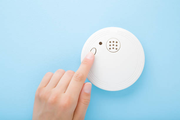 Young adult woman finger pushing button of new white plastic smoke alarm. Light blue table background. Pastel color. Closeup. Safety concept. stock photo